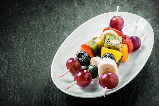 Colorful exotic fruit kebabs served on an oval dish with grapes, kiwifruit, orange, banana, blueberries, and strawberries on a dark background with copyspace