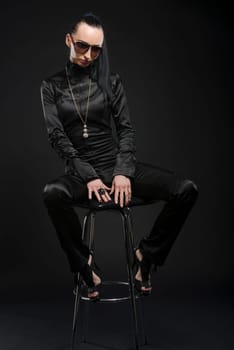 Full length shot of beautiful Caucasian woman wearing black bodysuit and sitting on a posing stool, against black background
