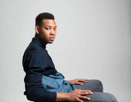 Portrait of handsome dark skinned male model sitting and wearing casual clothes against white background