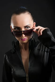 Headshot of beautiful Caucasian woman wearing black bodysuit and looking over sunglasses, against black background