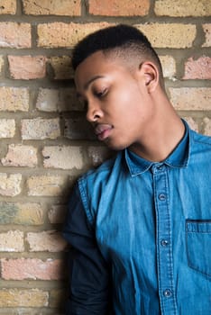 Portrait of handsome dark skinned male model wearing casual clothes and looking down, against brick wall