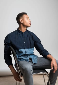 Profile portrait of handsome dark skinned male model sitting and wearing casual clothes against white background
