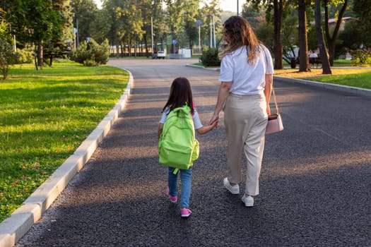 Mother and daughter walking together in park with backpack at sunset.