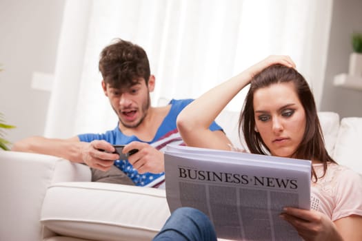 a couple in which he's playing videogames on his mobile phone and she's reading a business newspaper