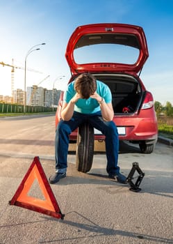 Portrait of sad man sitting on spare wheel near broken car