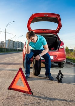 Depressed man sitting on spare wheel near broken car