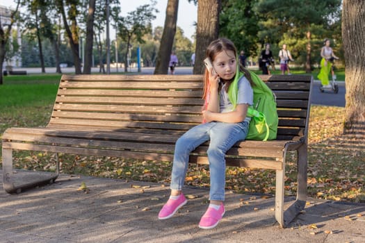 Young girl with backpack sitting on park bench in autumn sunlight.