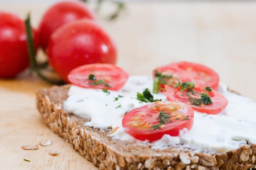 Close-up of a slice of bread coated with white cheese and three tomatoes lying behind it in a fuzzy background on a wooden cutting board  four slices of tomato and slightly chopped herbs as an additional bread topping are on top of