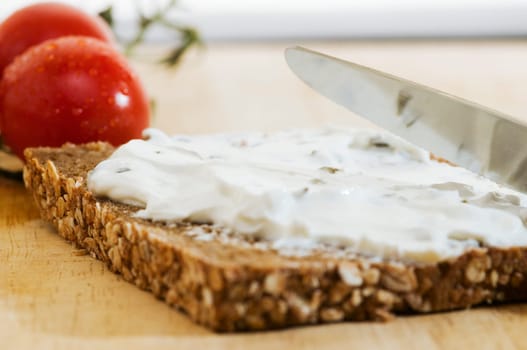Close-up of a slice of bread covered with white cheese and two tomatoes behind it on a wooden cutting board