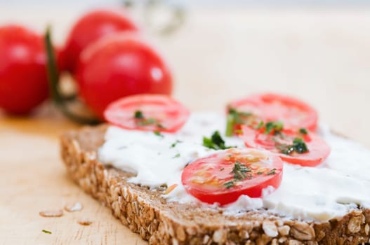 Close-up of a slice of bread coated with white cheese and three tomatoes lying behind it in a fuzzy background on a wooden cutting board  four slices of tomato and slightly chopped herbs as an additional bread topping are on top of