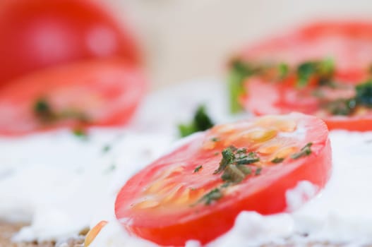 cut close-up of a slice of bread covered with white cheese,tomato slices and chopped herbs with a dissecting tomato lying on a wooden cutting board in the background  four slices of tomato and slightly chopped herbs as an additional bread topping are on top of
