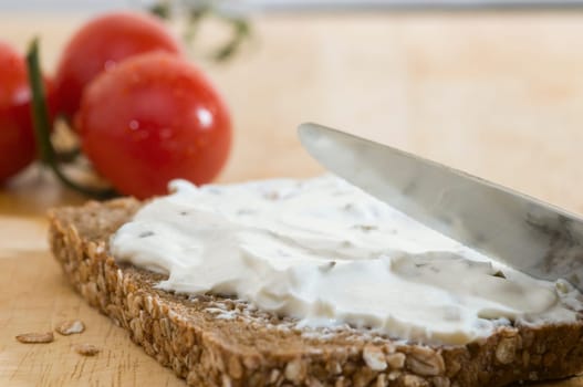 Close-up of a slice of bread coated with a knife of white cheese and two tomatoes lying behind it on a wooden cutting board