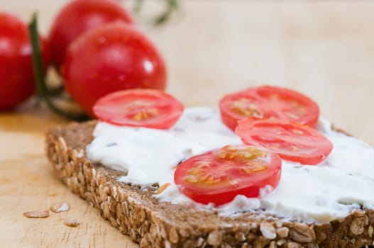Close-up of a slice of bread coated with white cheese and three tomatoes lying behind it on a wooden cutting board  four slices of tomato as an additional bread topping are on top of