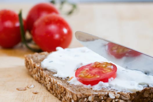 Close-up of a slice of bread coated with a knife of white cheese and two tomatoes lying behind it on a wooden cutting board  a tomato slice as an additional bread topping