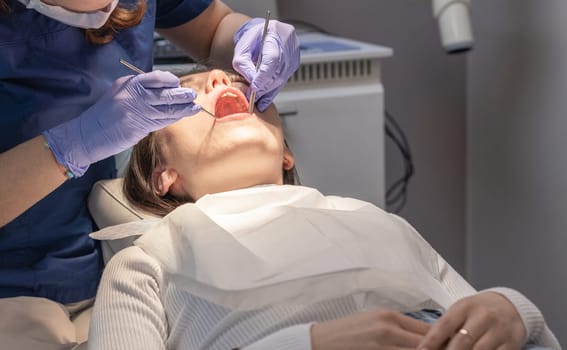 A dentist doctor in lilac gloves conducts a deep cleaning of the gums and teeth in the oral cavity using a tool to a young brunette girl patient lying in a chair, close-up side view. Concept, health and oral hygiene.
