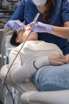A dentist doctor in a blue uniform and lilac gloves rinses the gums and teeth with water in the oral cavity using a tool to a young brunette girl patient lying in a chair, close-up side view. Concept, health and oral hygiene.