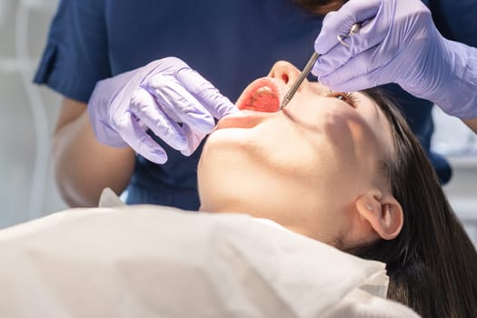 A dentist in a blue uniform and lilac gloves examines the gums and teeth in the oral cavity with a dental mirror to a young brunette girl patient lying in a chair, close-up view from below. Concept, health and oral hygiene.