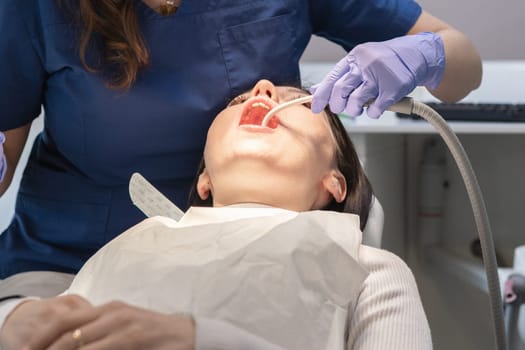 A dentist in a blue uniform and lilac gloves removes saliva with a dental saliva ejector into the mouth of a young brunette patient lying in a chair, close-up view from below. Concept, health and oral hygiene.
