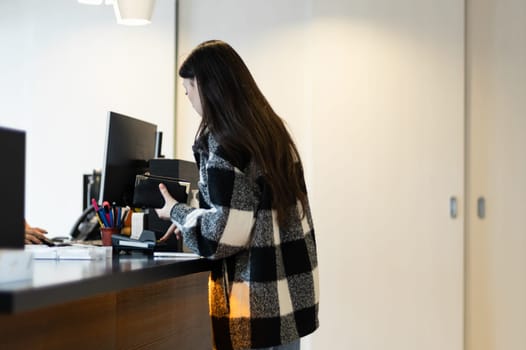 One young caucasian brunette woman with flowing hair in a plaid shirt is paying while standing at the reception desk in a dentistry, close-up side view.