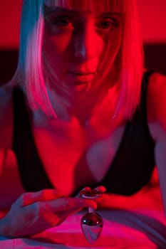 The blonde lies on the bed in red blue neon light and holds a steel butt plug