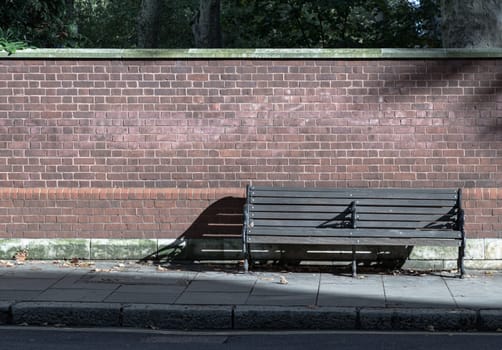 One empty bench made of wooden boards and black iron stands on sidewalk in the street with bricks wall background. Wooden vintage bench on walkway, Bench on pedestrian footpath, Seat and unwind area, Space for text, Selective focus.