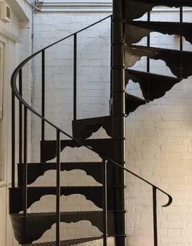 London, UK - Oct 24, 2023 - Vertical view of Black metal spiral staircase pattern with iron railing lead from the ground floor to the upper floor on the side of white brick wall. Ladder decoration exterior, Copy space, Selective focus.