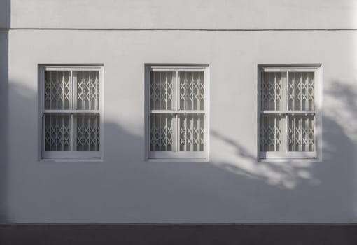 House exterior with triple wooden casement windows on white wall and white steel folding gates inside. Window with retractable grilles to prevent theft. The sunshine lights through the glass window frame, Space for text, Selective focus.