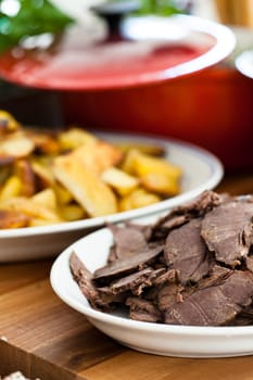 Closeup of slices of cooked beef on white serving plate with plate of thickly cut French fries in background