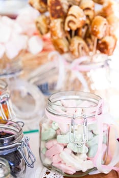 Closeup of pastel colored marshmallows in glass jar with decorative ribbon with small snack sized cinnamon buns on sticks in a glass jar in background