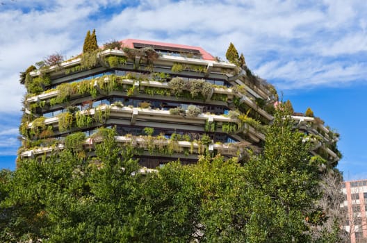 Facade of a building with greenery and flowers on a background of blue sky with clouds.