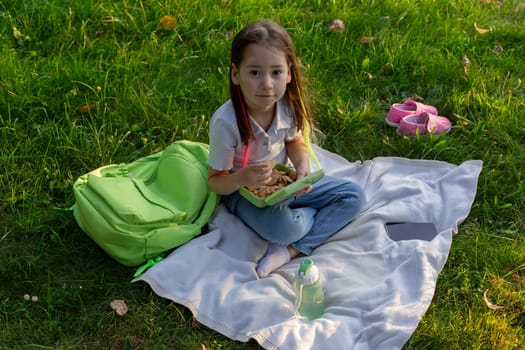 Young girl enjoying picnic on a blanket in the park with backpack and healthy lunch.
