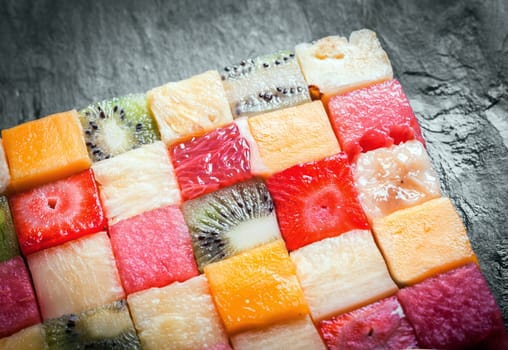 Cubed tropical fruit arranged in a decorative colorful square with a close up view showing the texture and pattern of the watermelon, kiwifruit, pineapple, strawberry and melon on a slate counter
