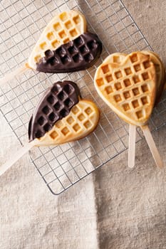 Heart-shaped freshly baked golden waffle lollipops, some with a chocolate coating on a wire mesh rack in the kitchen ready for a party or special occasion for a sweetheart or loved one