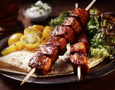 Healthy barbecued lean cubed pork kebabs served with a corn tortilla and fresh lettuce and tomato salad, close up view on a dark background