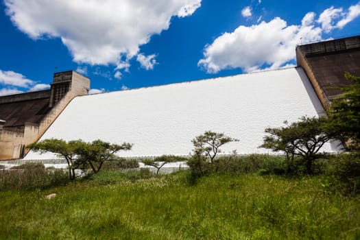 Dam filled to capacity and water flowing over the high wall with energy and power into summer green valley.