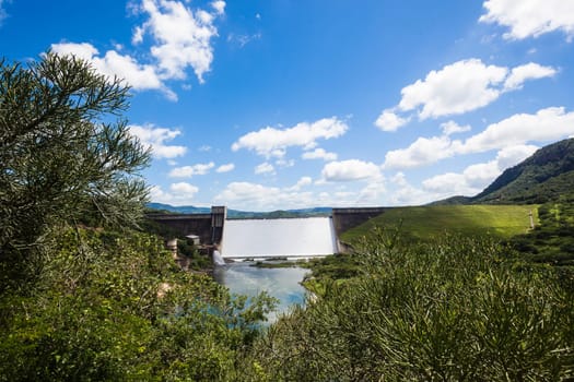 Dam filled to capacity and water flowing over the high wall with energy and power into summer green valley.
