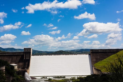Dam filled to capacity and water flowing over the high wall with energy and power into summer green valley.