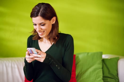 Beautiful caucasian girl chatting and text messaging on mobile phone sitting on sofa at home. Copy space