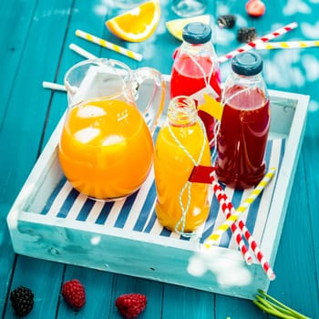 Bottles of freshly squeezed orange and berry juice standing on a wooden tray on a colorful turquoise blue picnic table in dappled summer sun