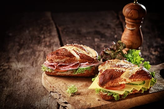 Crusty brown lye bread roll sandwiches with a cheese, salami, lettuce, tomato and cucumber filling on an old grunge board with a wooden pepper mill and copyspace