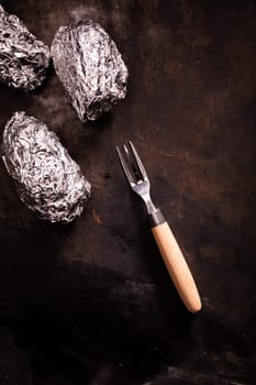 Barbecued foil wrapped potatoes still wrapped in their silver aluminium foil laying on an old slate surface with a fork and copyspace, overhead view
