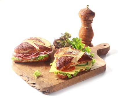 Traditional crusty brown German lye bread rolls, glazed with a lye solution before baking to produce the Maillard reaction, with savory cheese, salami and salad filling on a wooden board on white