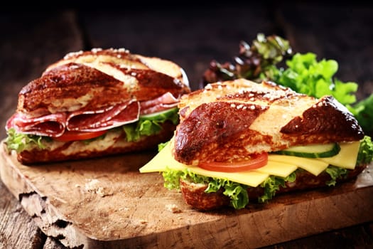 Crispy brown freshly baked lye bread rolls with cheese, spicy salami sausage, lettuce, tomato and cucumber on an old grunge rustic wooden chopping board