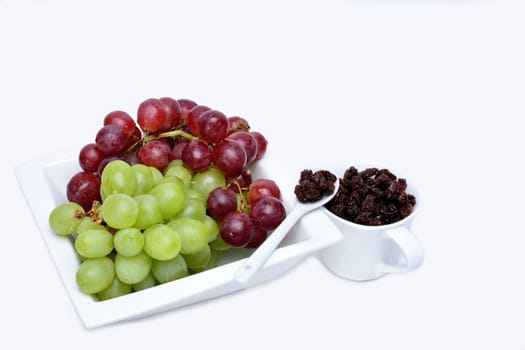 Bunch of red and green grapes on a white ceramic saucer and a spoon and cup of raisins