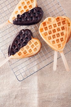Romantic heart shaped waffle lollipops with a crisp golden wafer dipped on one side in dark chocolate cooling on a wire tray in a kitchen for Valentines or an anniversary