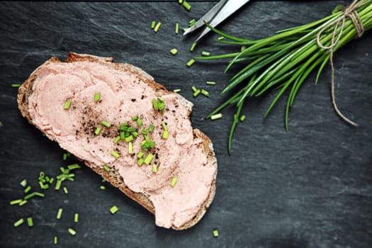 Preparing a tasty smoked teewurst sandwich on a slice of rye bread with chopped chives and black pepper, overhead view on a slate kitchen counter with copyspace