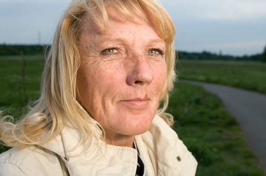 Head-and-shoulder portrait of a blonde mature woman in bright jacket looking at the camera on a green lawn with footpath