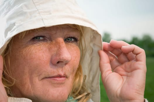 side head portrait of a blonde mature woman in bright jacket with hood on a green meadow looking contentedly into the camera Fingers of the left hand on the side of the hood