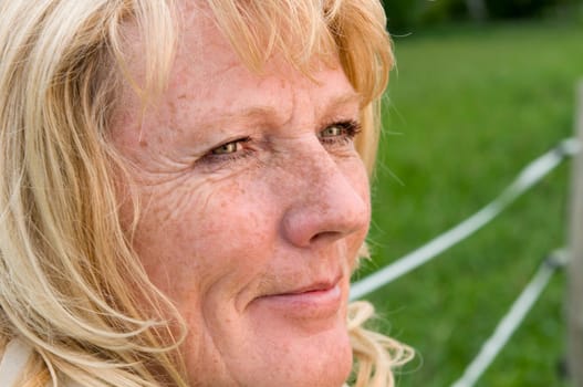 Side close-up of a blonde mature woman on the fence on a green meadow thoughtful and contentedlooking looking into the light
