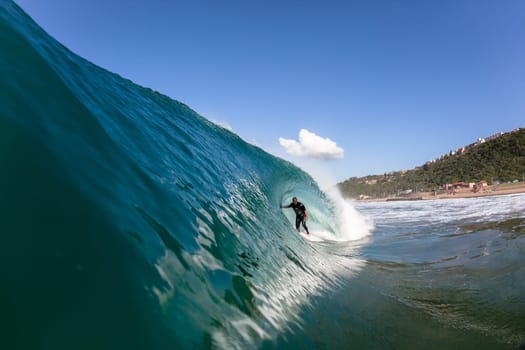 Surfer tube rides hollow blue wave wall upright round crashing breaking onto reef beach shallows, a water swimming photo perspective